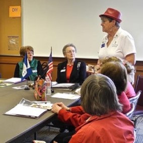This photo shows women sitting around a table listening to a woman in a red hat who is standing.