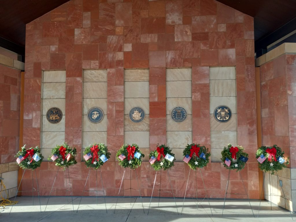 This photo shows a row of wreaths on stands, each with a U.S. flag and a red bow, in front of a marble wall with the emblems of the five U.S. military services.