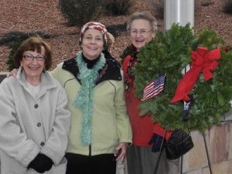 This photo shows three ladies posing with a large green wreath with a red bow before they participate in Wreaths Across America.